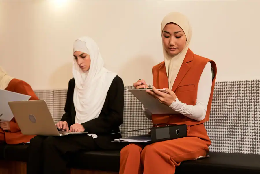 Two hijabi women working in a modern office setting, using a laptop and writing notes on clipboard.