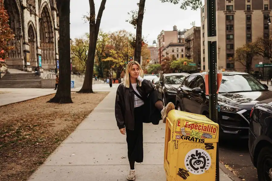 A woman strolls by a cathedral with fall foliage and urban backdrop.